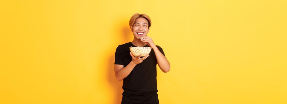 Carefree happy asian guy with blond hair, watching comedy, laughing and eating popcorn, standing yellow background.