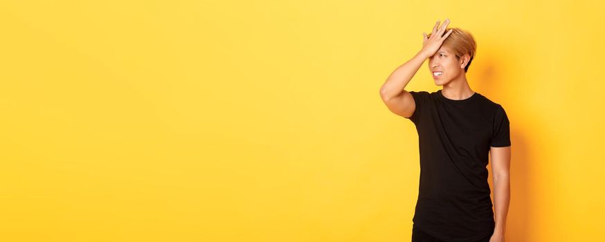 Upset and troubled asian guy snap forehead forgetful, standing over yellow background.