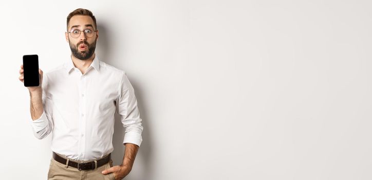 Surprised handsome manager in glasses, looking curious and showing mobile screen, standing over white background.