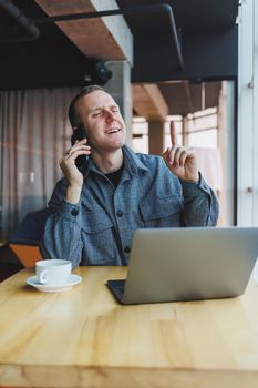 Smiling male entrepreneur in casual wear sitting at table with laptop and talking on smartphone in modern workspace with large window during day