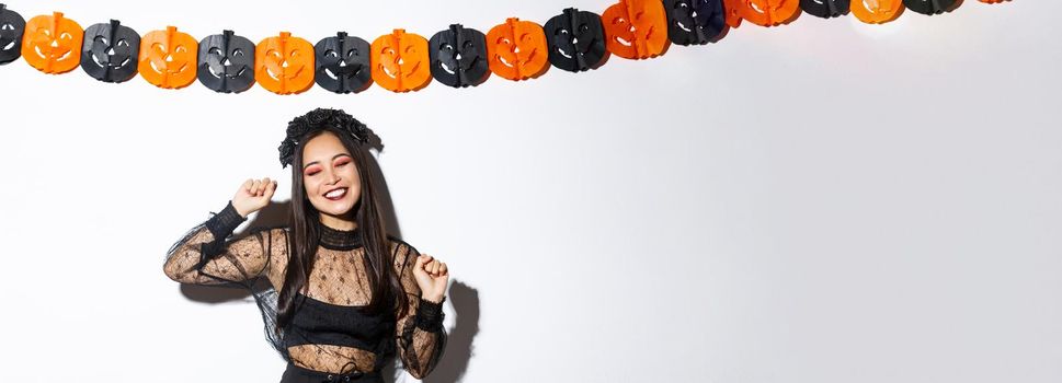 Carefree smiling asian woman in witch costume enjoying halloween party, dancing and rejoicing, standing over white background with pumpkin decoration.