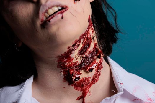 Halloween monster having bloody wounds on neck, posing in studio over blue background. Undead eerie zombie eating brain and being wounded, having ugly dirty scars and scratches with blood. Close up.
