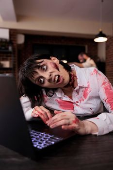 Scary horrific zombie using laptop at desk, undead corpse trying to work on computer in startup office. Creepy aggressive brain eating monster looking terrifying and horrible, sinister danger.