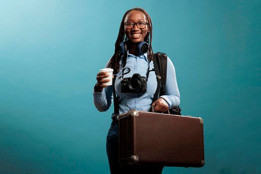 Happy traveler with holiday suitcase and DSLR camera smiling optimistic at camera while getting ready for vacation. Smiling tourist with photo device and wireless headphones waiting to go on journey.