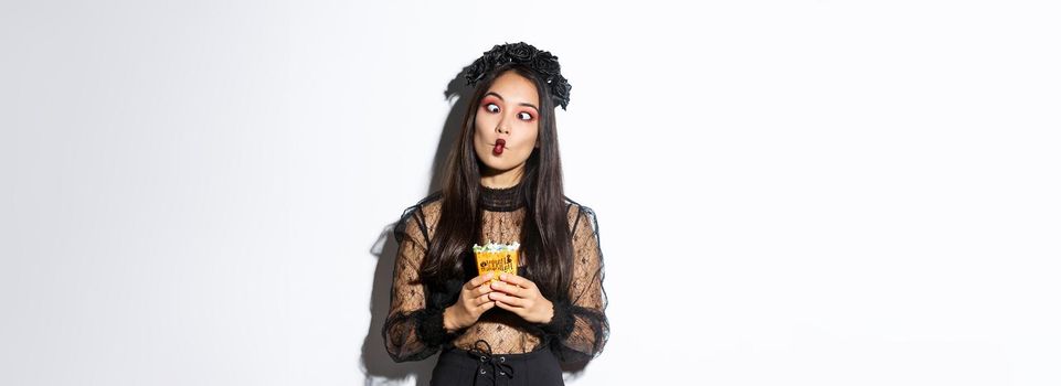 Image of funny carefree girl celebrating halloween in witch costume, making silly faces and holding treats, standing over white background.