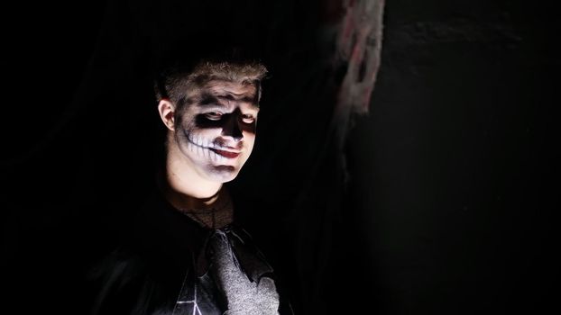 Halloween party, night, frightening portrait of a man in the twilight, in the rays of light. man with a terrible make-up in a black witch costume. High quality photo