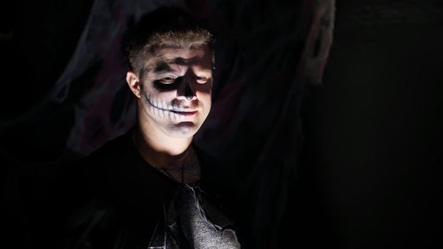Halloween party, night, frightening portrait of a man in the twilight, in the rays of light. man with a terrible make-up in a black witch costume. High quality photo