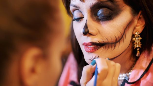 Halloween party, make-up artist draws a terrible makeup on the face of a brunette woman for a Halloween party. in the background, the scenery in the style of Halloween is seen. High quality photo