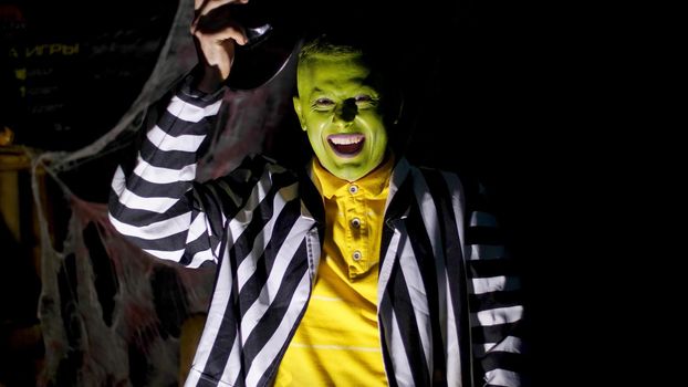 Halloween party, night, twilight, in the rays of light, a man with a terrible make-up, with a green face lifts his hat, laughs eerily and runs away. Behind the background is a spiderweb. High quality photo