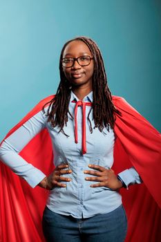 Joyful happy smiling young adult person posing as justice defender on blue background. Proud superhero woman with superpower abilities wearing mighty hero cloak while smiling confident at camera
