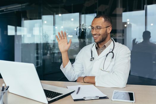 Happy hispanic doctor smiling and talking on video call man in white coat and stethoscope using laptop for remote online consultation smiling and waving hello.