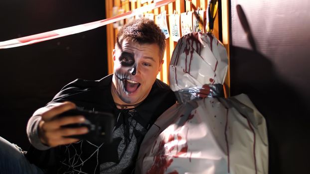 Halloween party, night, twilight, in the rays of light, a man with a terrible makeup does selfie with a corpse wrapped in oilcloth, the corpse is smeared with blood. High quality photo
