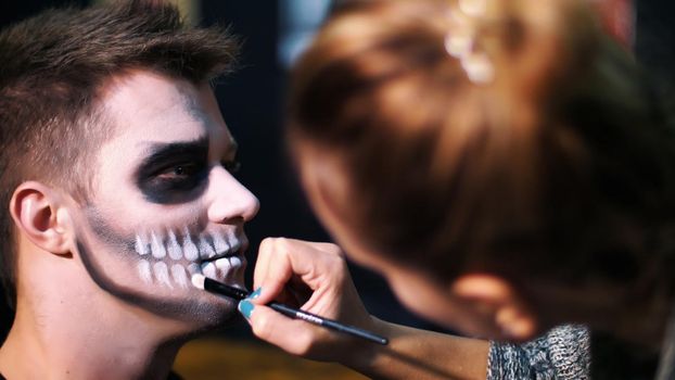 Halloween party, close-up, make-up artist draws a terrible makeup on the face of a man for a Halloween party. in the background the scenery in the style of Halloween is seen. High quality photo