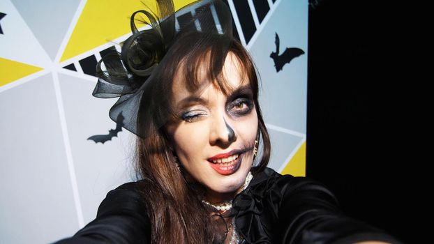Halloween party, night, frightening portrait of a woman with a terrible makeup in a black witch suit, croaks in front of the camera. High quality photo