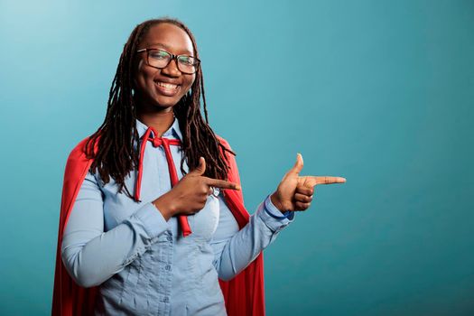 Studio shot of cheerful and optimistic justice defender woman wearing red cape while pointing fingers to right on blue background. Selfless and brave young adult superhero person posing at camera.