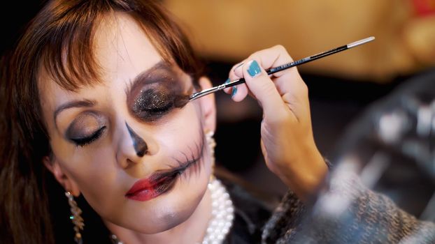 Halloween party, close-up, make-up artist draws a terrible makeup on the face of a brunette woman for a Halloween party. the scenery in the style of Halloween is seen. High quality photo
