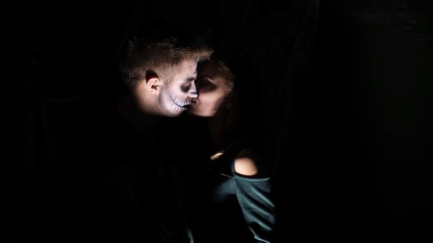 Halloween party, night, frightening man in the twilight, in the rays of light, with a terrible make-up in a black witch costume. demon kisses the girl. the werewolf's kiss. High quality photo