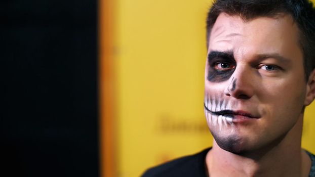 Halloween party, close-up, make-up artist draws a terrible makeup on the face of a man for a Halloween party. in the background the scenery in the style of Halloween is seen. High quality photo