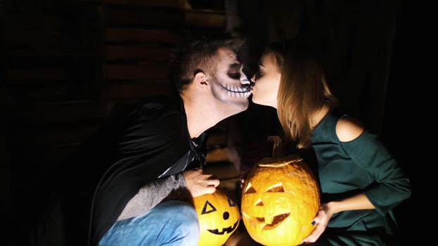 date in style of Halloween party, night, twilight, in the rays of light, guy with a girl dressed in costumes and with a terrible makeup are holding two large Halloween pumpkins, kissing. High quality photo