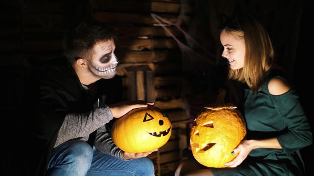 date in style of Halloween party, night, twilight, in the rays of light, guy with a girl dressed in costumes and with a terrible makeup are holding two large Halloween pumpkins. High quality photo