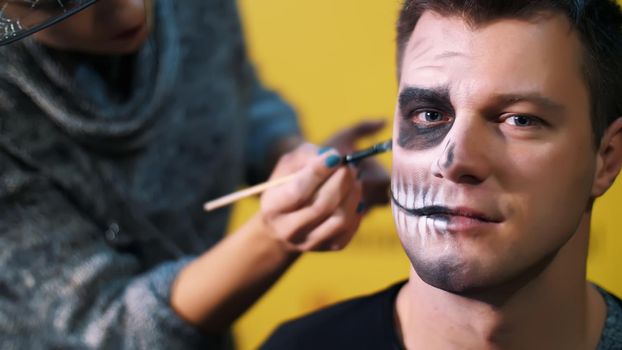 Halloween party, close-up, make-up artist draws a terrible makeup on the face of a man for a Halloween party. in the background the scenery in the style of Halloween is seen. High quality photo