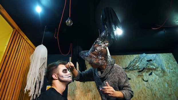 Halloween party, make-up artist draws a terrible makeup on the face of a man for a Halloween party. in the background, the scenery in the style of Halloween is seen. High quality photo