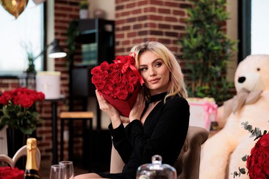 Woman in black dress holding valentines day red roses, receiving luxury presents. Young blonde girl with flower bouquet sitting in living room, filled with expensive romantic gifts