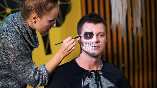 Halloween party, make-up artist draws a terrible makeup on the face of a man for a Halloween party. in the background, the scenery in the style of Halloween is seen. High quality photo