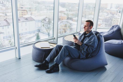 Young male student in a casual suit resting in a comfortable pouf while studying, an attractive freelancer in a suit works remotely