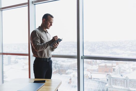 A young businessman stands at a panoramic window in a skyscraper and makes notes in his notebook with a pen. A man in a shirt and trousers of European appearance