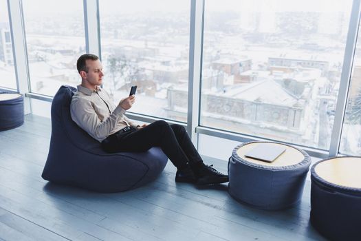 Successful male freelancer sitting in modern workspace and checking notifications on mobile phone in casual clothes. Remote work.