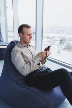 Successful male freelancer sitting in modern workspace and checking notifications on mobile phone in casual clothes. Remote work.
