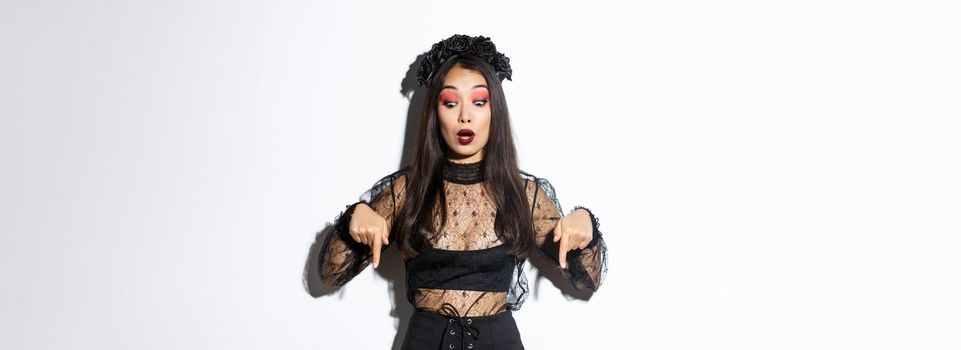 Surprised beatiful asian woman in black gothic dress and wreath, pointing fingers down and looking startled at something about halloween or trick or treat, standing over white background.