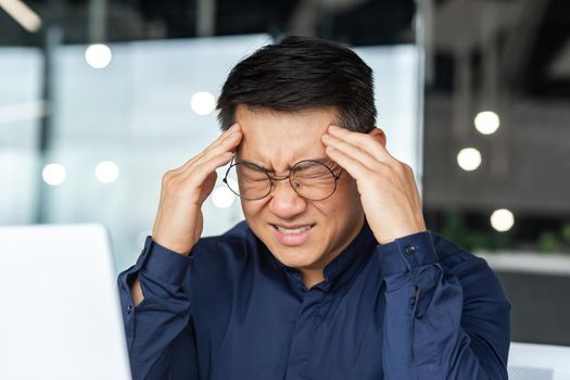 Close-up photo of sick Asian businessman having severe headache, male boss in glasses working inside modern office building, holding hands on head massaging, overtired at work.