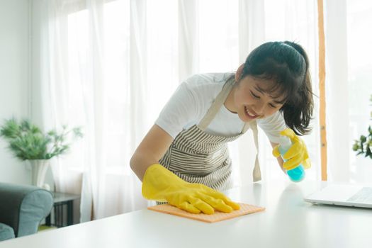 Young woman happily cleaning, wiping, disinfecting table surface using cleaning spray and sponge and wearing yellow protective gloves at home. Woman doing housechores with enjoyment.