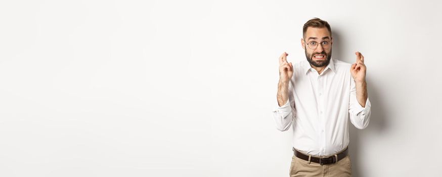 Worried man making a wish, cross fingers and hope for relish, standing over white background.