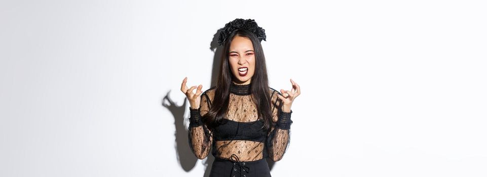 Image of woman in halloween costume looking like angry evil witch, cursing and clenching fists mad. Female in black dress and wreath trick or treating, standing white background.