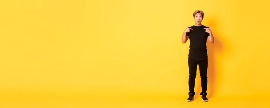 Full-length of surprised attractive korean blond guy in black outfit pointing at himself impressed, yellow background.