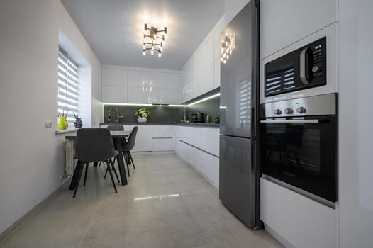 Luxurious modern trendy white and grey kitchen interior after renovation, with granite counter top and marble floor, dining table next to window