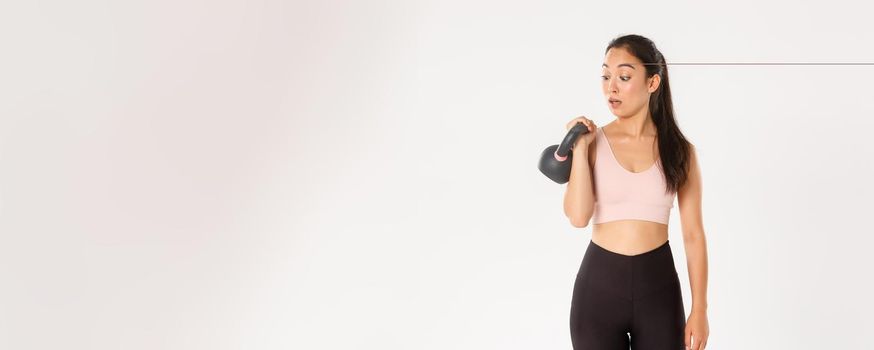 Sport, wellbeing and active lifestyle concept. Portrait of cute brunette asian fitness girl, sign up bodybuilding classes at gym, surprised with weight of kettlebell, standing over white background.