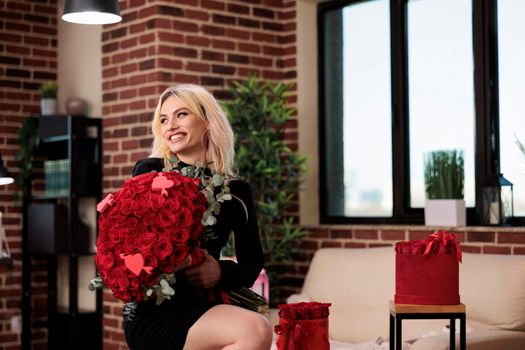 Woman posing with luxury red roses bouquet, laughing, sitting on bar stool in living room, medium shot. Beautiful blonde girl with valentines day expensive gift, romantic date.