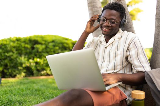 Young black man listening to music with headphones and using laptop outdoors. College student doing homework on campus. Copy space. Education and technology concepts.