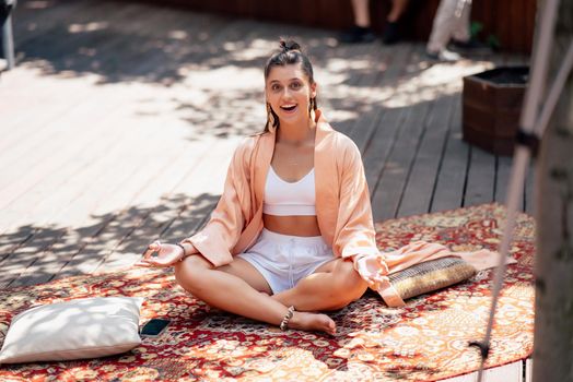 Young woman is practising yoga sitting on a wooden podium in the garden in the sunny morning