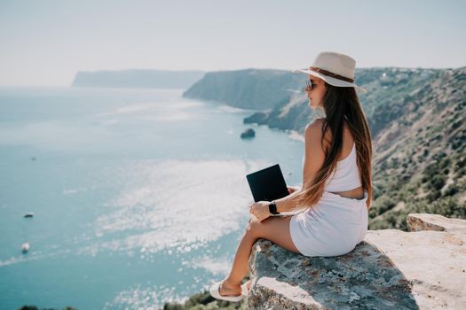 Successful business woman in yellow hat working on laptop by the sea. Pretty lady typing on computer at summer day outdoors. Freelance, travel and holidays concept.