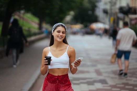 Cheerful woman in the street drinking morning coffee and use her smartphone. Young woman using phone on the street.