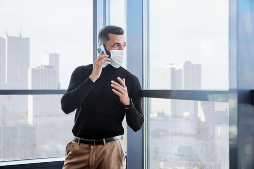 man in a protective mask talking on a smartphone standing near the window.