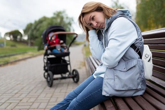 young woman mother of postpartum depression sits with built-in feelings in the park on a bench next to a baby stroller.