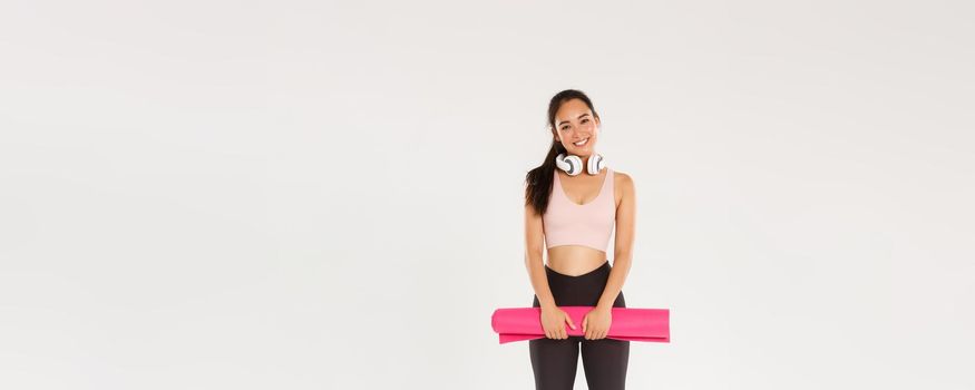 Full length of cute asian girl like fitness, holding rubber mat for exercises or yoga classes, standing in sportswear looking happy after productive training in gym, good workout, white background.