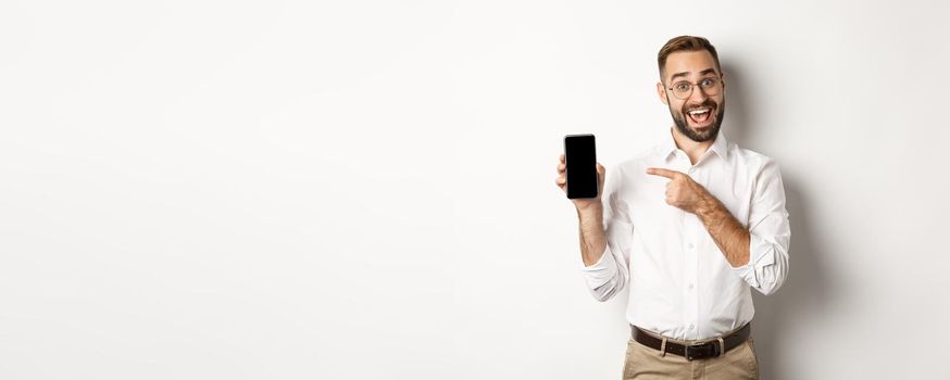 Excited handsome guy showing mobile phone, pointing finger at screen and smiling, standing against white background.
