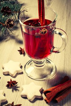 Glass of Christmas mulled wine with cinnamon, star anise and cloves on wooden background with white biscuits, natural fir tree branches and cones. Color toning effect.